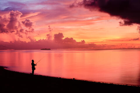 The sky turns colorful as the sun sets on Saipan as a silhouetted man surf fishes. 