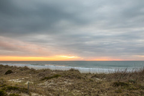 The sun rises over the ocean and beams through clouds warming the dunes and seas oats