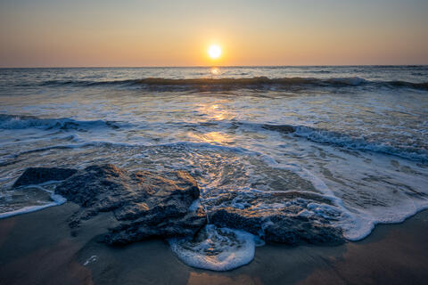 The sun rises over the Atlantic Ocean at folly beach, sc, at lighthouse inlet heritage preserve with rocks in the foreground.