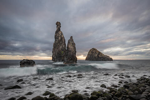 Miradouro da Ribeira da Janela is seen at dawn with the basalt rocks visible in the foreground as the sky lights up with color as the sun rises. 