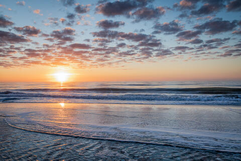 The Sun rises over the Atlantic ocean at dawn lighting up the clouds and the waves which break on the beach.