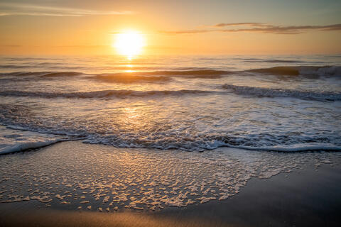 The Sun rises over the Atlantic ocean at dawn lighting up the clouds and the waves which break on the beach. Lack of wind shows as there is a clam sea. 
