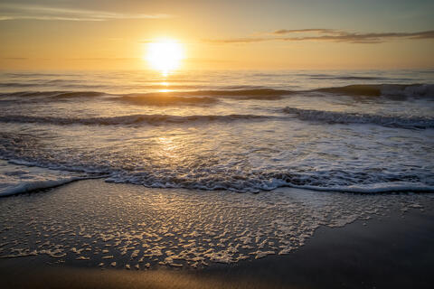 The Sun rises over the Atlantic ocean at dawn lighting up the clouds and the waves which break on the beach. Lack of wind shows as there is a clam sea. 